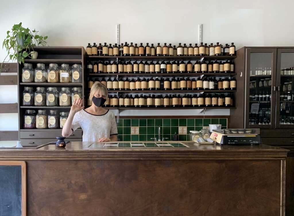 blonde woman waving hello behind front desk with botanical tincture displays